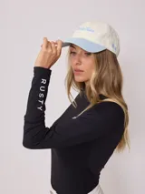 Gorra de lona color crema con visera celeste y bordado frontal con el logo de la marca Rusty en celeste.