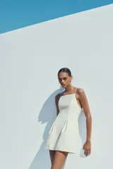 Vestido corto blanco de tejido elástico, con escote recto, tirantes finos ajustables, detalle de pliegues y cierre con cremallera oculta en la costura.