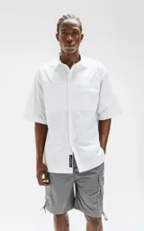 Camisa de manga corta, color blanco, confeccionada en popelín de algodón con silueta oversize. Posee dos bolsillos frontales, uno con solapa y otro con ojal y botón.