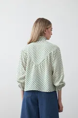 Camisa de viscosa blanca con estampa de lunares negros. Presenta cuello alto con volado y botonadura frontal, mangas largas con puños abullonados y frunces bajo el busto.