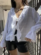 Camisa blanca traslúcida con diseño a cuadros, escote en V con lazo, mangas largas abullonadas con puños elásticos y corte holgado.