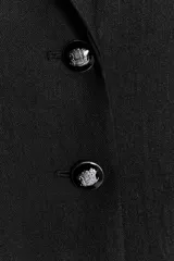 Blazer larga negra con cuello solapa, manga larga y bolsillos de vivo en el delantero. Cierre frontal con dos botones plateados.