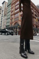 Vestido midi de manga larga, color marrón oscuro, confeccionado en tejido de encaje o punto con patrón floral sutil. Presenta cuello perkins y drapeado asimétrico en el torso con frunces laterales.