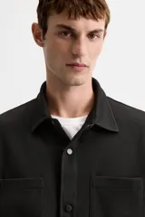 Sobrecamisa negra de tejido con elasticidad, cuello solapa y manga larga acabada en puño con botón a presión. Tiene bolsillos de plastrón en el pecho y cierre frontal con botones a presión.