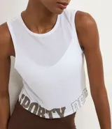 Musculosa deportiva corta (crop top) blanca, con cuello redondo y sisas amplias. Presenta un panel frontal de tela transparente/mesh sobre el busto y la palabra "SPORTY" estampada en color marrón en el bajo.