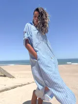 Vestido camisero largo celeste con rayas verticales blancas, cuello camisero, mangas cortas con trabilla y botones, cierre frontal con botones y bolsillos laterales.