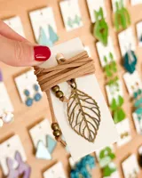 Collar tipo choker con tiento de gamuza marrón claro, enrollado en una tarjeta de presentación. Incluye un dije metálico calado con forma de hoja en tono bronce envejecido, adornado con cuentas redondas del mismo tono.