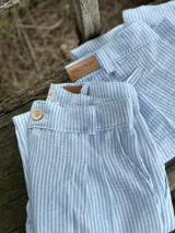 Short de lino celeste a rayas verticales blancas, con botón de madera y etiqueta de cuero con el logo de la marca.