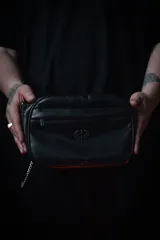 Bandolera negra de cuero sintético con correa ancha ajustable y desmontable. Cuenta con cierre superior, bolsillo frontal con tiracierre customizado, forro interno y cuatro bolsillos en total (dos exteriores y dos interiores).