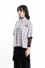 Camisa unisex de manga corta con estampado a cuadros en tonos verde, negro, blanco y morado. Presenta un corte boxy y un estampado en serigrafía en el pecho.