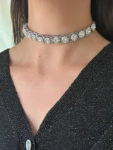 Gargantilla choker color plateado, con tiento trenzado simil gamuza metalizada y apliques de flores con strass. Se cierra con cadena de acero quirúrgico y gancho ajustable.