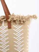 Bolso tipo tote color beige con diseño geométrico en blanco y flecos en la parte superior. Tiene doble asa de hombro marrón.