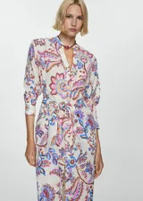Vestido midi de corte camisero con estampado floral en tonos rosa, azul y marrón sobre fondo blanco. Tiene cuello mao, mangas 3/4 con puños abotonados, cierre frontal con botones y lazo decorativo en la cintura.