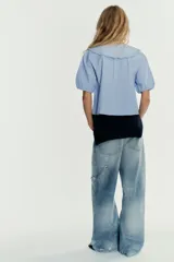 Camisa celeste a rayas verticales blancas, de corte recto, con cuello bobo con bordados y escote pico con lazada. Tiene mangas cortas abullonadas con elástico y cierre frontal con botones.