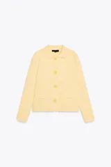 Chaqueta de punto color amarillo pálido, con cuello solapa y manga larga. Tiene bolsillos delanteros de plastrón y cierre frontal con botones con forma de flor.