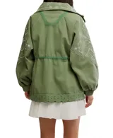 Chaqueta estilo parka o anorak de algodón color verde militar, con capucha y cordón ajustable en la cintura. Presenta parches de tela con bordados blancos de estilo paisley o floral en la parte delantera, mangas y capucha. El ruedo inferior tiene un borde festoneado con ojales.