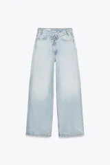Pantalón de jean celeste de tiro alto, corte wide leg y largo hasta el tobillo. Presenta un diseño asimétrico en la cintura con cierre frontal cruzado con cremallera y botones metálicos.