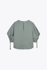 Blusa de cuello redondo color verde, confeccionada en hilatura con mezcla de lyocell. Manga corta abullonada ajustable con lazadas. Bajo redondeado.