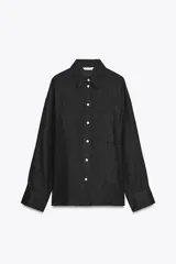 Camisa amplia color negro confeccionada con hilatura de lino. Tiene cuello solapa, manga larga, bolsillo de plastrón en el delantero, bajo asimétrico y cierre frontal con botones.