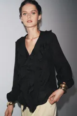 Camisa color rosa pálido confeccionada en tejido ligero con lyocell. Presenta escote en V, cierre frontal con botones, mangas largas abullonadas con puños elásticos y detalle de volantes en el frente.