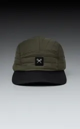 Gorra de estilo 5 paneles, color verde militar, con corona acolchada y visera negra. Presenta un pequeño parche negro con un logo blanco en forma de tijeras cruzadas.