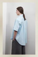 Camisa celeste de popelina de algodón, corte amplio, con cuello clásico, manga corta y bolsillo frontal. Presenta canesú con pliegue central en la espalda.