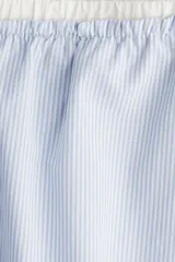 Short de popelina de algodón a rayas verticales celestes y blancas. Tiene cintura elástica con cordón ajustable, bolsillos laterales y aberturas laterales cortas en el bajo.