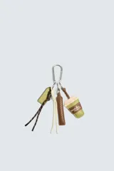 Llavero con mosquetón metálico plateado y varios charms colgantes: una etiqueta rectangular verde oliva, una correa trenzada marrón, un cordón trenzado blanco y amarillo, una correa trenzada marrón y un dije de vaso de bebida verde con la inscripción "matcha moment".