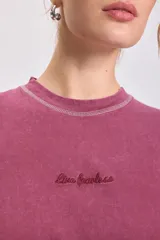 Remera de manga corta color bordó con efecto lavado, corte holgado y costuras externas visibles en blanco en cuello, mangas y ruedo. Presenta un pequeño bordado cursivo en el pecho.