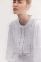 Blusa blanca de algodón con cuello mao, pechera con jaretas e inserciones de encaje, botones frontales, canesú fruncido en la espalda y mangas largas abullonadas con pinzas y abertura con botón en los puños.