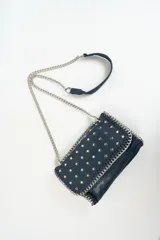 Cartera bandolera azul con tachas plateadas en forma de estrella, correa de cadena plateada y asa corta de cuero.