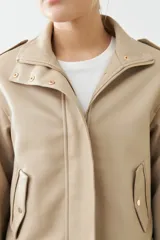 Chaqueta corta color beige de cuero sintético, con cuello alto, cierre frontal con botones a presión y bolsillos laterales con solapa.