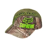 Gorro de gabardina verde con visera y panel frontal con estampado de camuflaje de ramas y hojas. Logotipo de Fox Racing y Supreme bordados en verde flúor en el panel frontal.