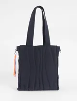 Bolso tipo shopper acolchado color beige claro, con costuras verticales onduladas en hilo azul. Posee dos asas de hombro y un pequeño accesorio colgante con cordones de colores en uno de los laterales. Presenta un logo cuadrado negro en el centro frontal.