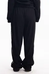 Pantalón de jogging negro con textura acanalada, corte relajado y cintura elástica. Presenta una franja gris con borde blanco a lo largo de la pierna y logo bordado en el muslo.
