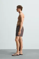 Bañador corto de hombre con estampado de rayas verticales en marrón, blanco y azul. Cuenta con cintura elástica ajustable con cordón, bolsillos laterales y trasero.