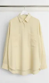 Camisa color amarillo claro, de corte holgado, con cuello clásico, cierre frontal con botones, mangas largas y dos bolsillos de parche en el pecho.