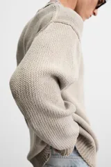 Jersey de punto color beige, de corte holgado, con cuello redondo y manga larga. Tiene efecto lavado y rotos en toda la prenda.