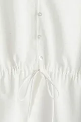 Blusa blanca de algodón con textura de lunares. Tiene escote redondo con lazo, botones en el frente, mangas tres cuartos abullonadas, cordón ajustable en la cintura y peplum.