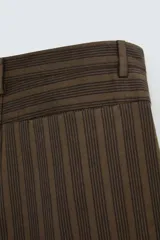 Pantalón marrón con finas rayas verticales negras, de corte amplio tipo balloon fit, con bolsillos laterales y traseros ocultos. Cierre frontal con cremallera y botonadura cruzada.