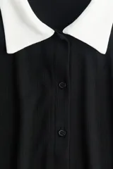 Camisa negra de punto acanalado con cuello blanco, manga corta y botonadura frontal.