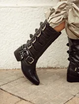 Bota de cuero vacuno color negro, de caña alta y punta fina, con detalles de tiras con hebillas plateadas. El interior está forrado en cuero vacuno. Tiene un fondo de doble vira de madera.