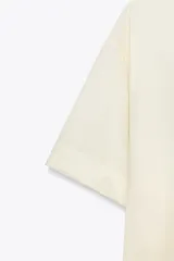 Camisa color crema de cuello solapa y manga corta con vuelta. Tiene un bolsillo de parche en el delantero, bajo asimétrico con aberturas laterales y cierre frontal con botones.