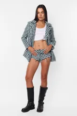 Conjunto de blazer y short de tiro alto con estampado pata de gallo en tonos verde y blanco. El blazer es de corte oversize, con solapas y cierre cruzado con botones. El short es de corte ajustado con terminaciones al corte con costura de seguridad. Se completa el look con un top blanco y botas de cuero negras.