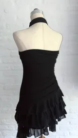 Vestido negro, sin mangas, con escote asimétrico y detalle de lazo o corbatín largo que cae desde el hombro. El cuerpo es ajustado y fruncido, y la falda corta tiene volados superpuestos.