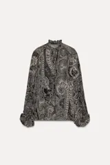 Camisa de viscosa con estampado paisley en blanco y negro. Cuello redondo con escote pico ajustable con cordones, mangas largas con elástico y volantes en los puños, bajo asimétrico y cierre frontal con botones forrados.