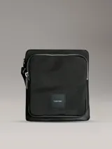 Bolso tipo reportero o bandolera Calvin Klein de color negro, con cuerpo principal de tejido texturizado y detalles en material liso en los bordes. Presenta un bolsillo exterior con cierre de cremallera en la parte superior y un parche cuadrado con el logo de Calvin Klein en la parte frontal inferior.