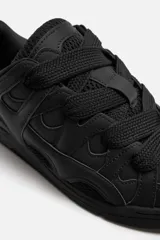 Zapatilla deportiva tipo skate color negro. Parte superior fabricada en combinación de piezas y materiales. Detalle de cordones maxi y suela con acabado irregular.
