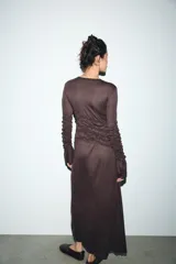 Vestido midi color marrón oscuro, con cuello redondo y manga larga. Presenta frunces diagonales en la parte superior delantera y en las mangas, y un bajo asimétrico.