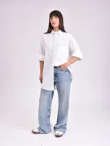 Camisa blanca de corte amplio, con cuello clásico, mangas 3/4 con puños arremangados y bolsillo de parche en el pecho.